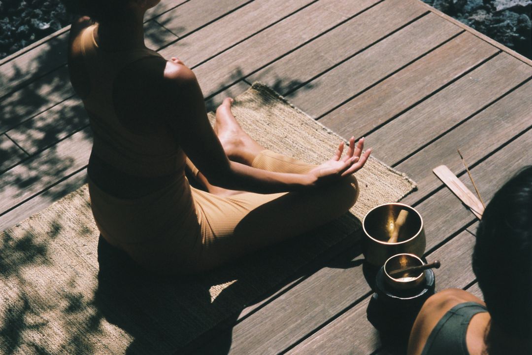 A woman meditating on a wooden deck with  singing bowls next to her.