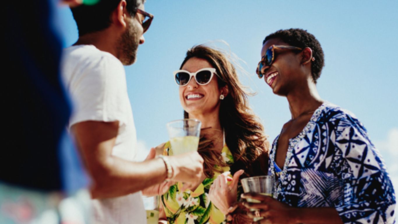 dos mujeres y un hombre con gafas ríen y beben en la playa 