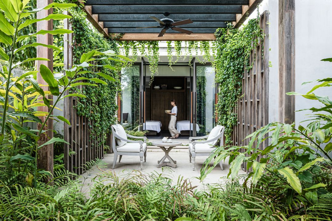 A spa seating area with chairs, a table, and plants, featuring a person walking in the background.