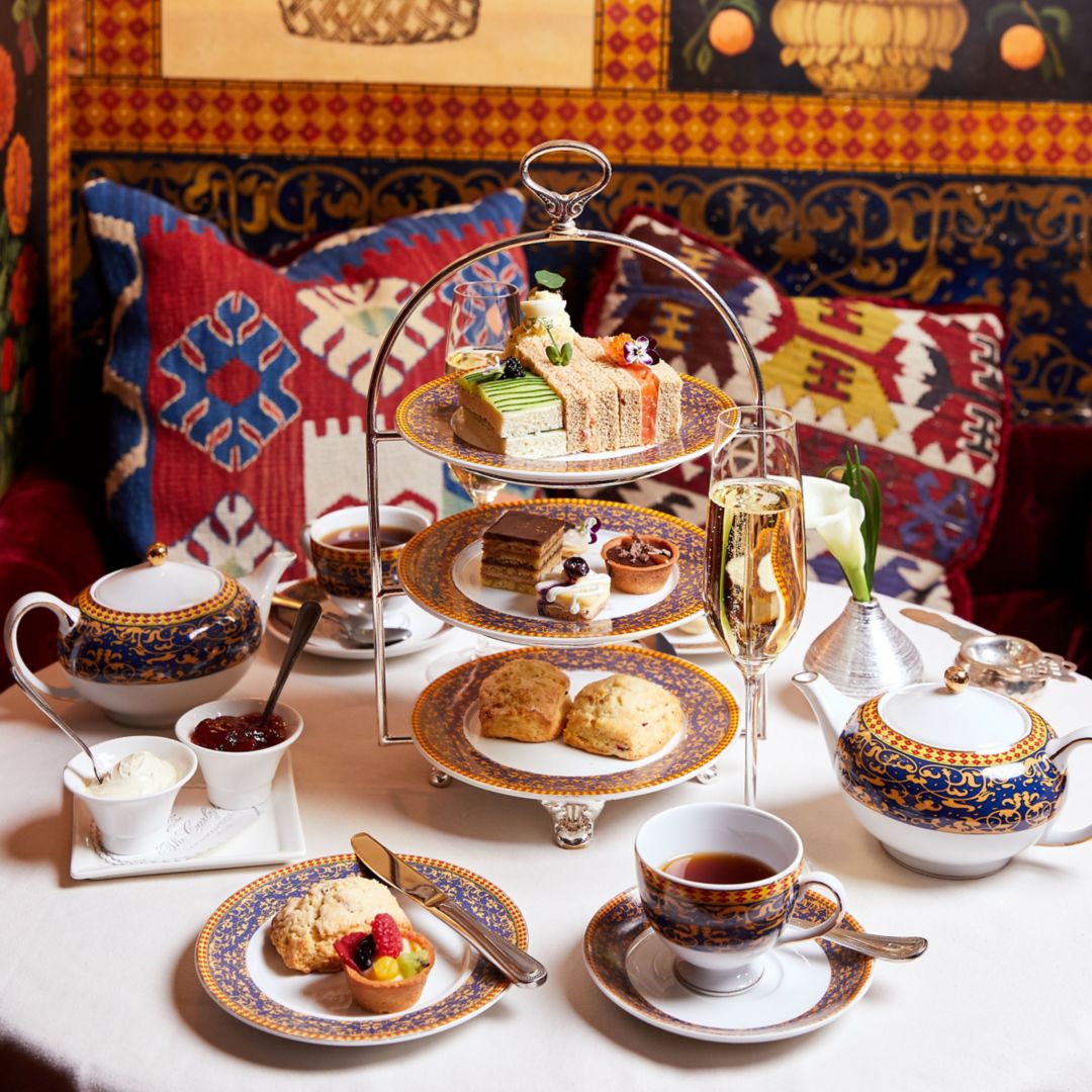 A table set for afternoon tea with a tiered stand of sandwiches, scones, and cakes, accented by patterned cushions.