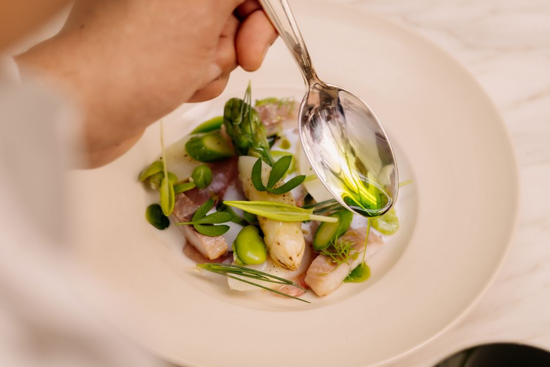A hand drizzles olive oil over a dish with  vegetables on a white plate.
