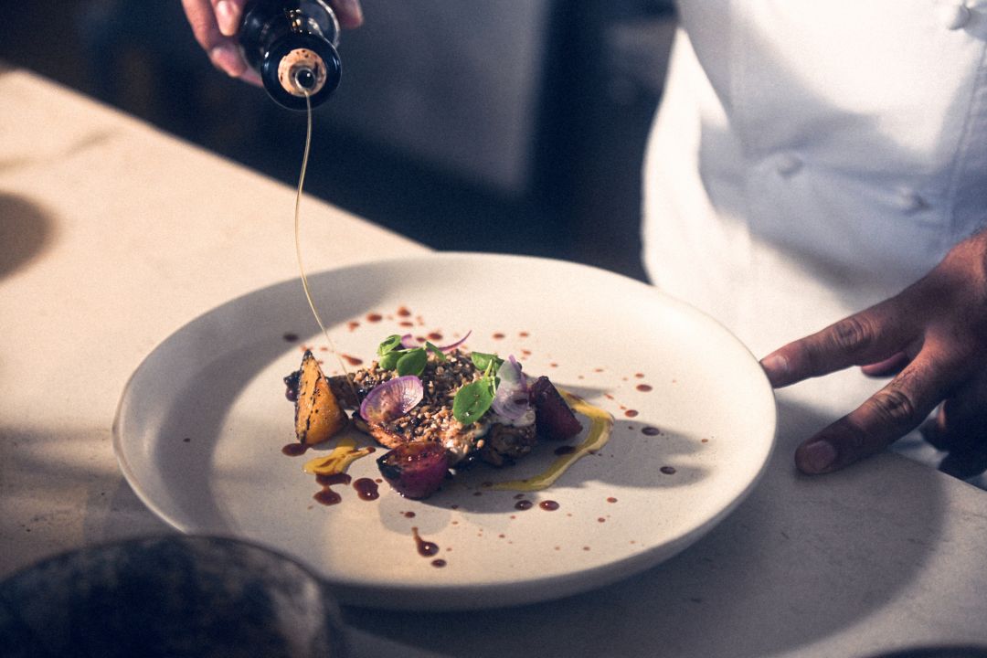 A chef drizzles sauce over a plated dish with  vegetables and garnishes.