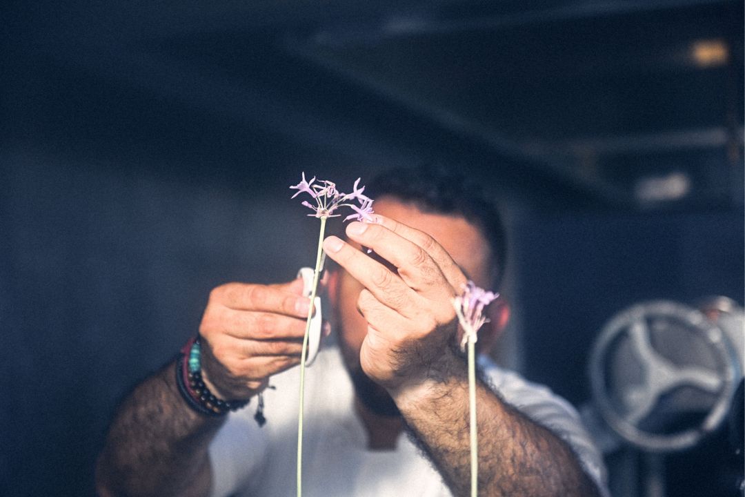 A chef holding a small purple flower in front of  his face, with a blurred background.