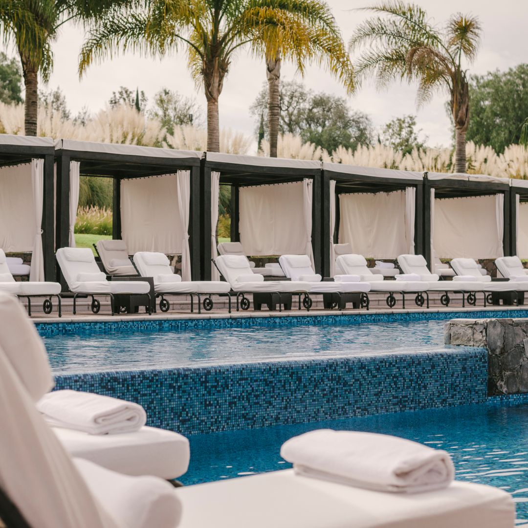 A hotel pool area with lounge chairs under canopies, palm trees, and rolled towels.