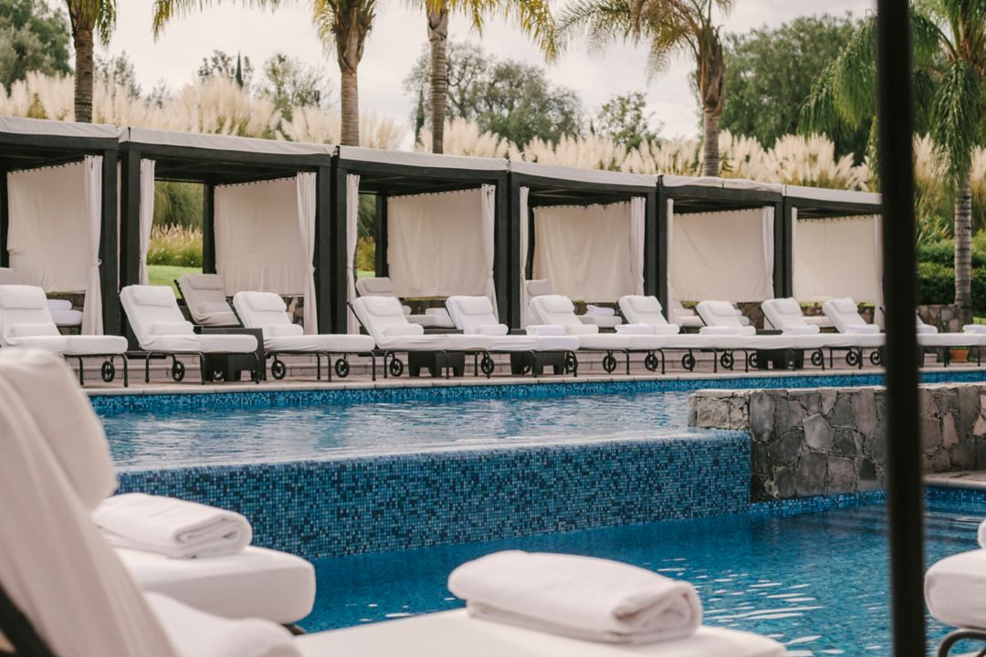 A hotel pool area with lounge chairs under canopies, palm trees, and rolled towels.