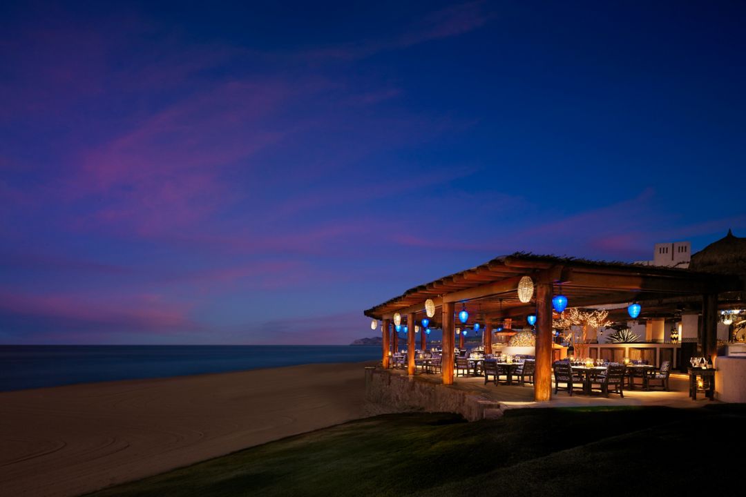 A beachfront restaurant at dusk, with a thatched roof, warm lighting, and a view of the ocean.