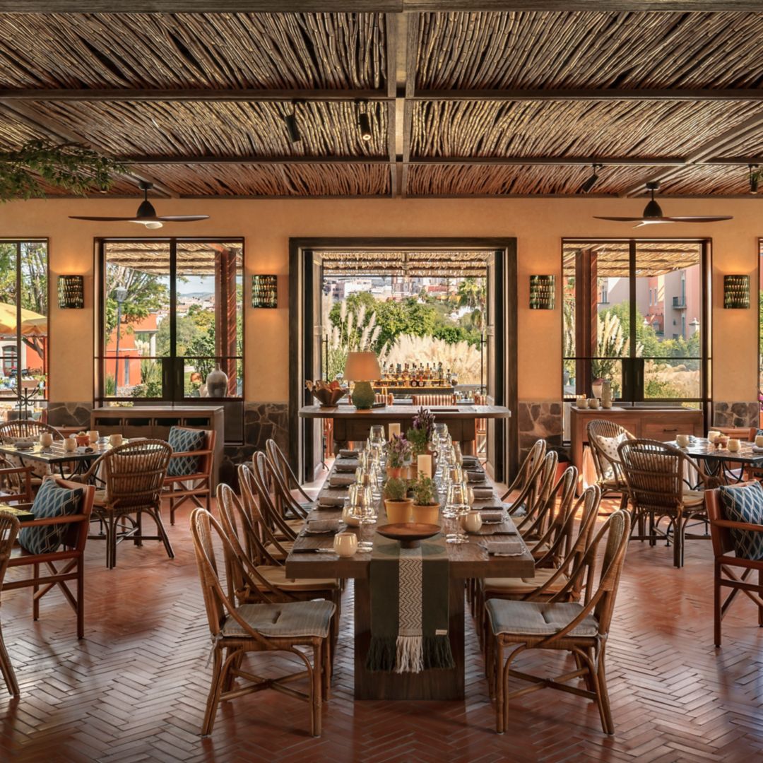 A restaurant in San Miguel de Allende with tables, chairs, a bar, and large windows offering outdoor views.