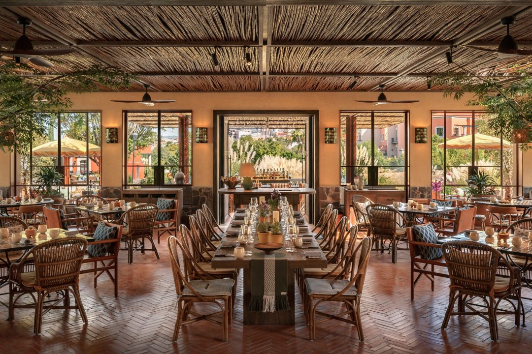 A restaurant in San Miguel de Allende with tables, chairs, a bar, and large windows offering outdoor views.