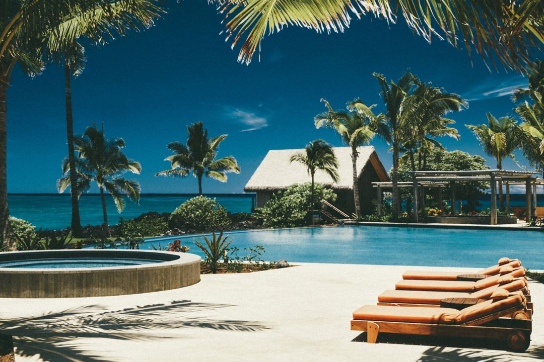 Luxury resort infinity pool with ocean views, surrounded by palm trees, sun loungers, and a thatched-roof villa.