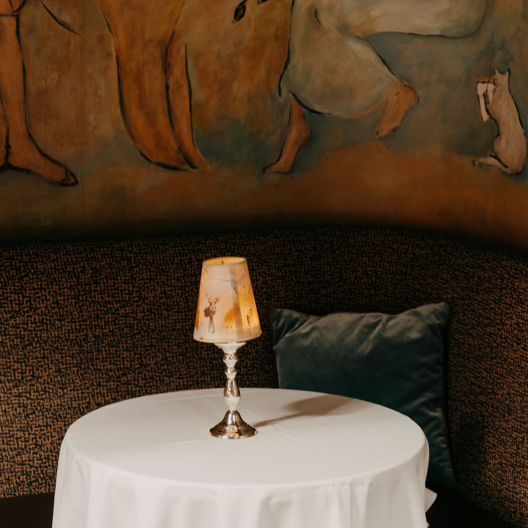 A mural with figures and a bird, above a table with glassware and a decorative lamp.