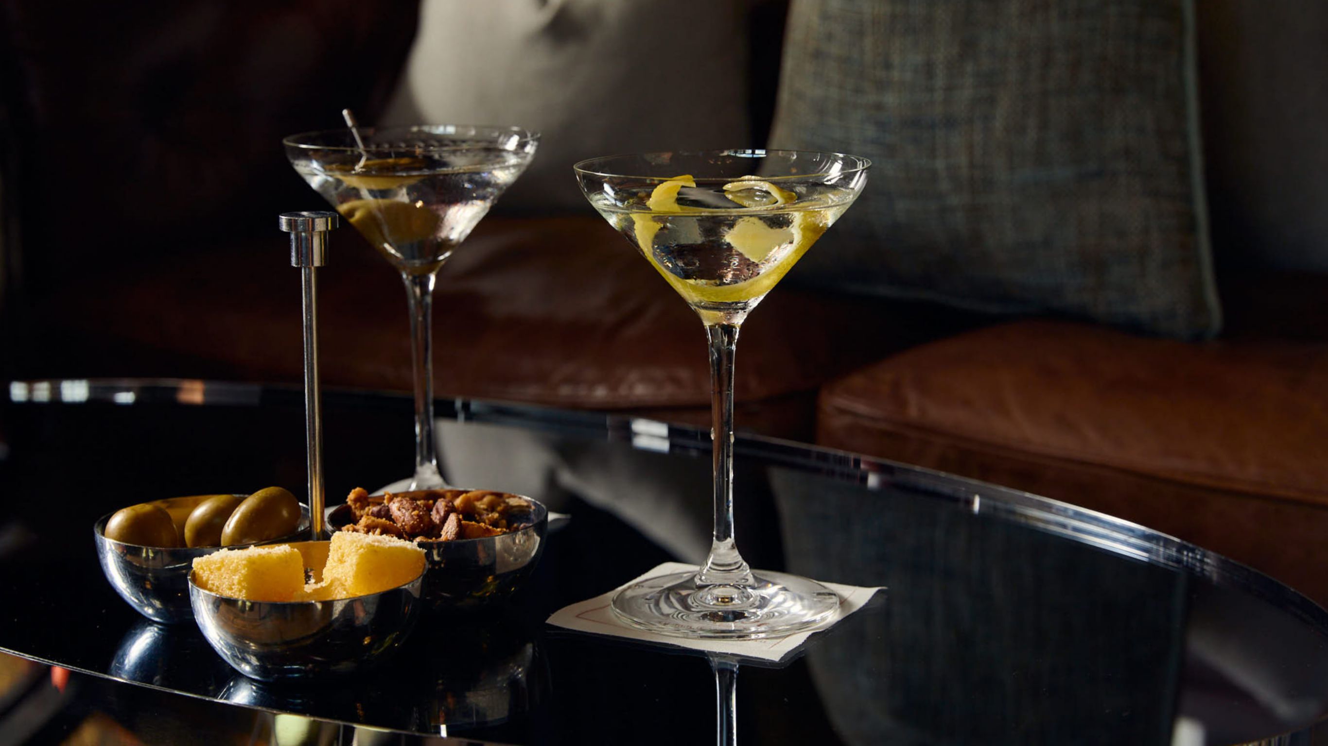 Two martini glasses with olives and lemon twists, accompanied by a bowl of olives and nuts on a table.