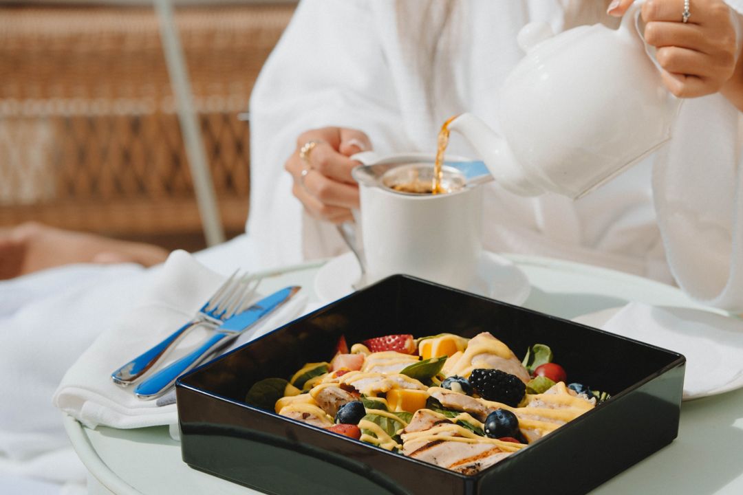 A woman, dressed in a white robe, relaxes as she enjoys a salad and pours herself some tea.