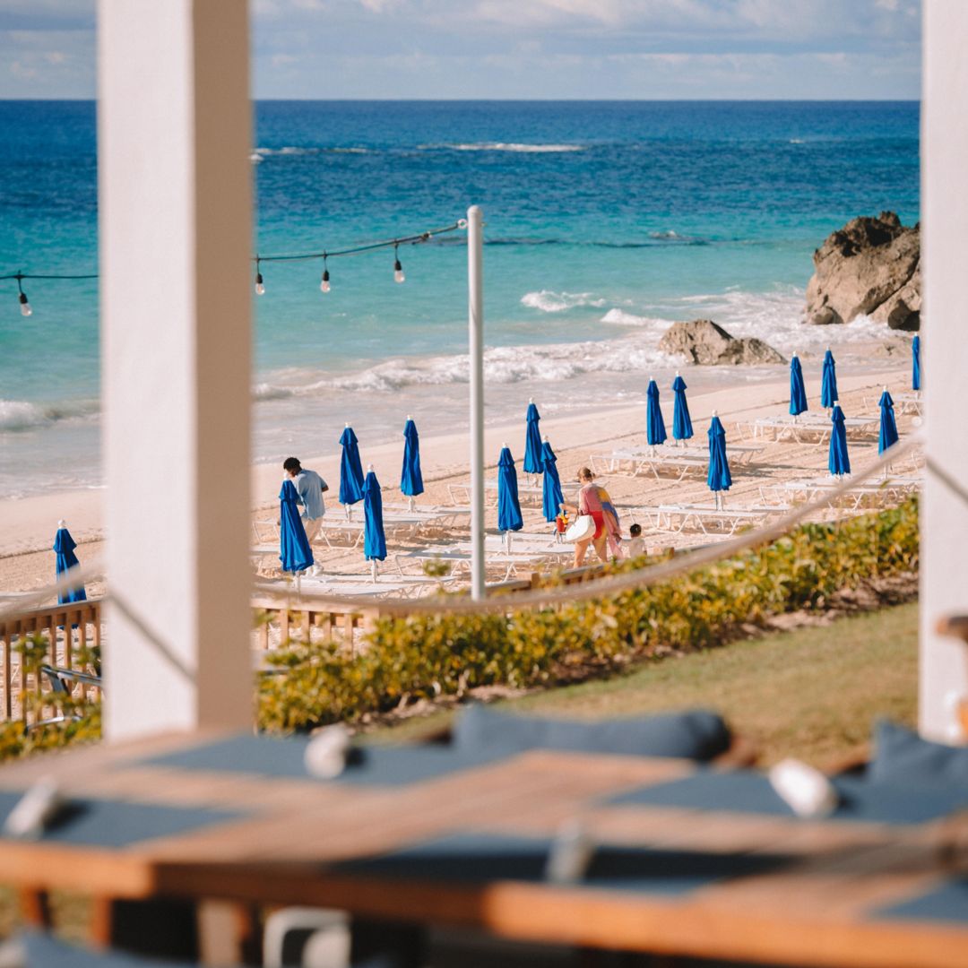 A table at a seaside restaurant in Bermuda overlooks a family on the beach, passing by chairs and unopened umbrellas.