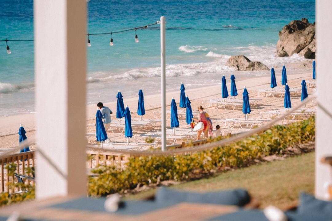 A table at a seaside restaurant in Bermuda overlooks a family on the beach, passing by chairs and unopened umbrellas.