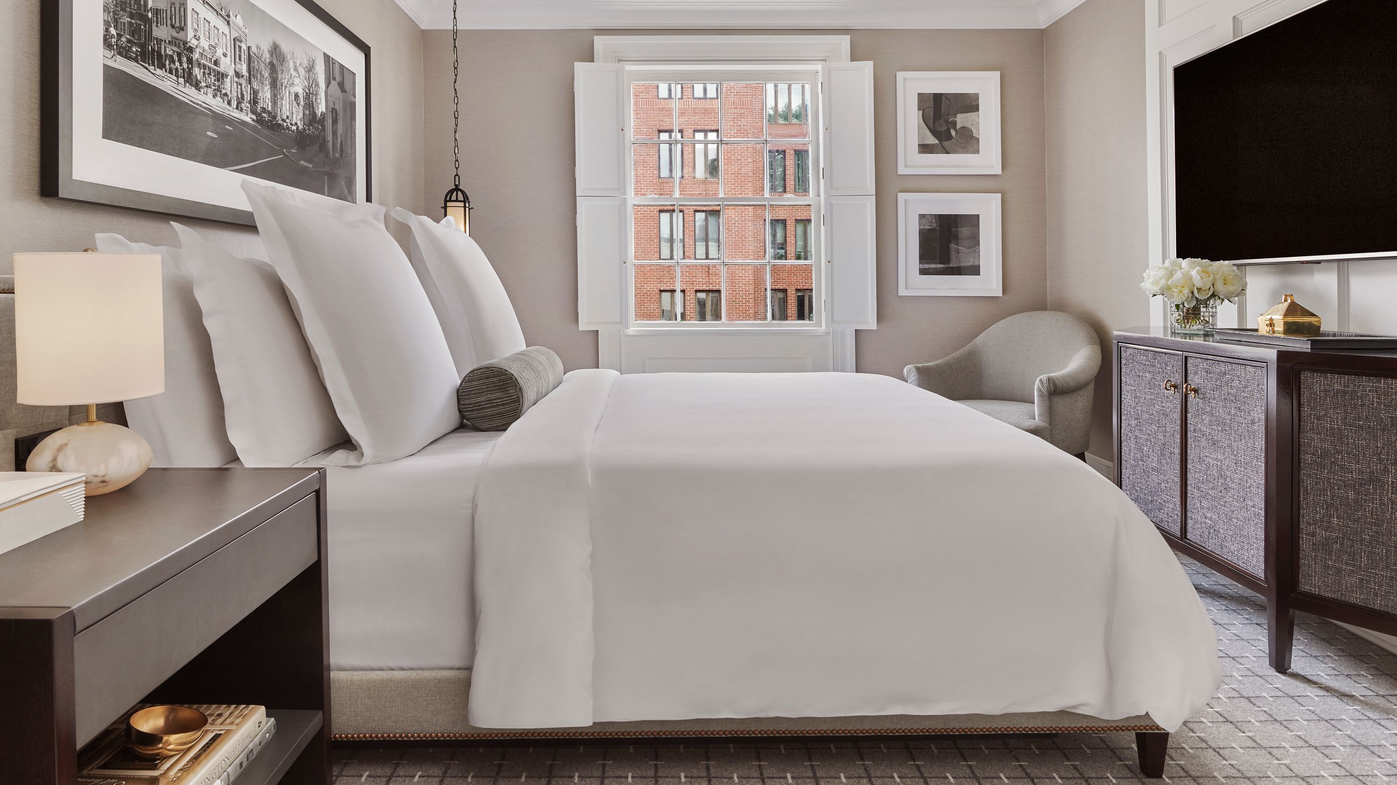 A bedroom with a bed, nightstand, lamp, chair, dresser, and a window with a view of buildings.