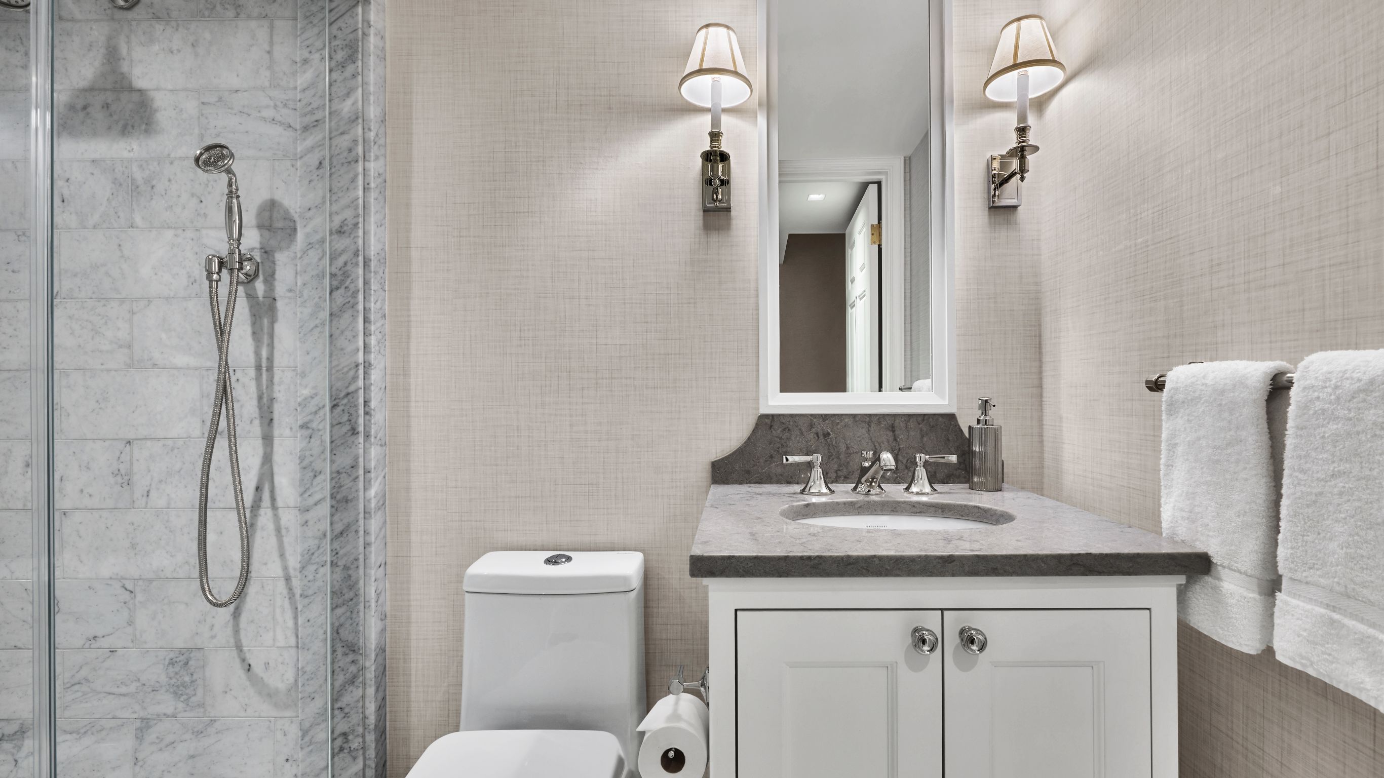 A bathroom with a sink, mirror, toilet, shower, wall lamps, and towels hanging on a rack.