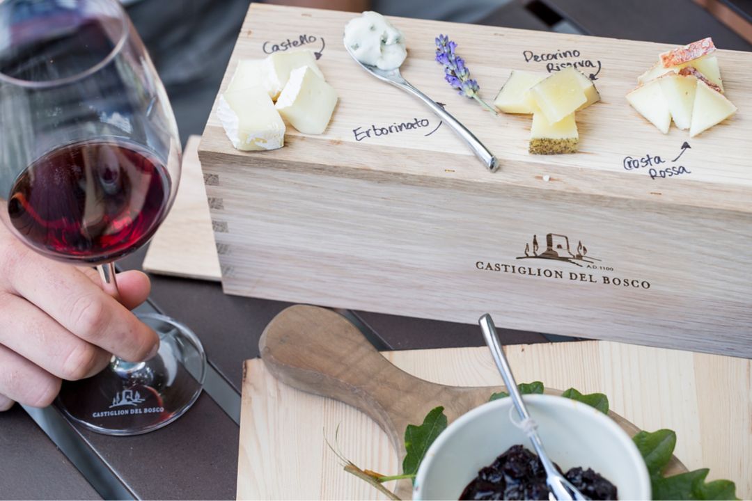 Wooden board with various cheeses and a spoon, alongside a glass of red wine.