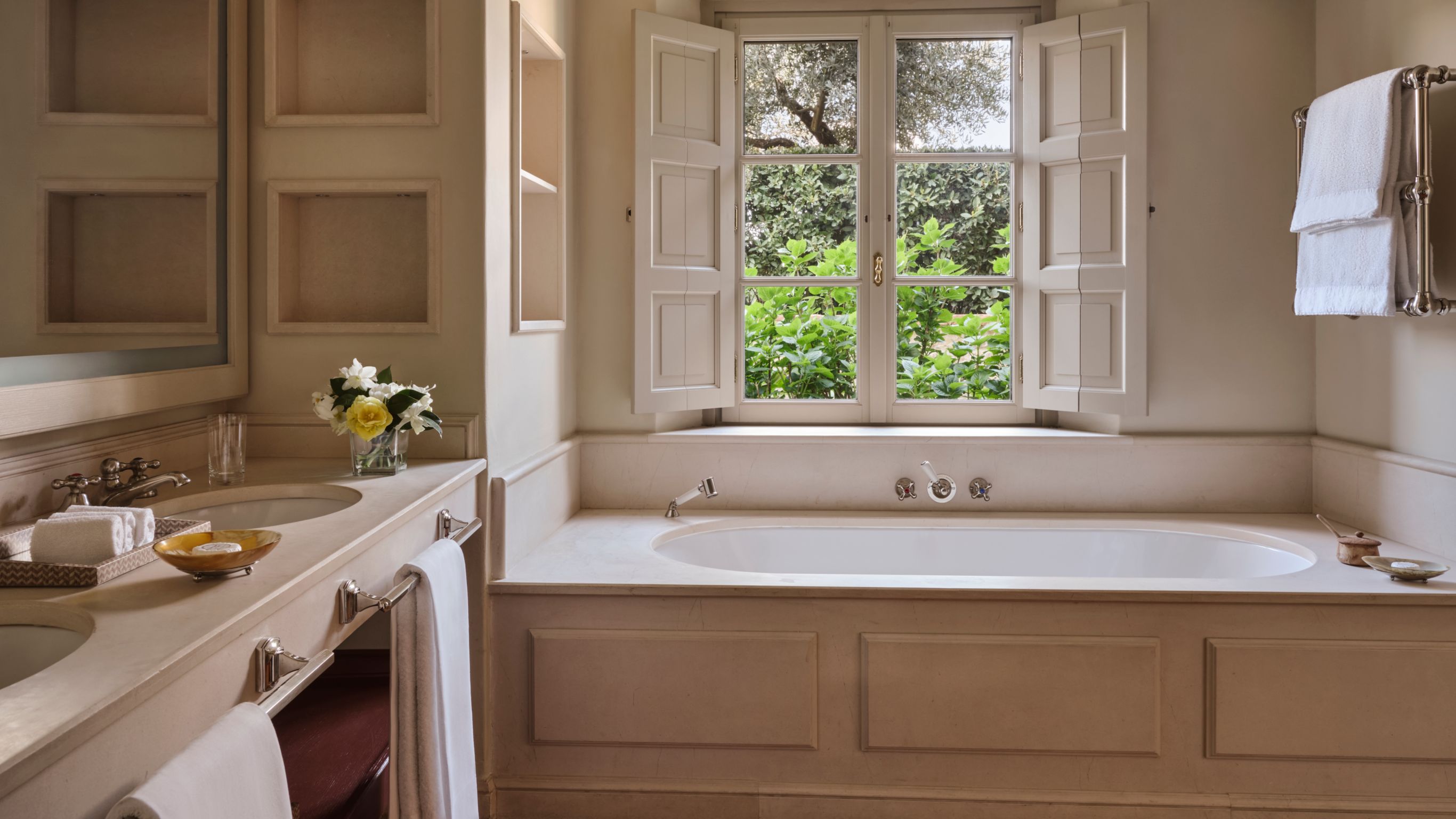 An elegant bathroom in a villa with a large bathtub under a window, vanity with flowers, and towels on a rack