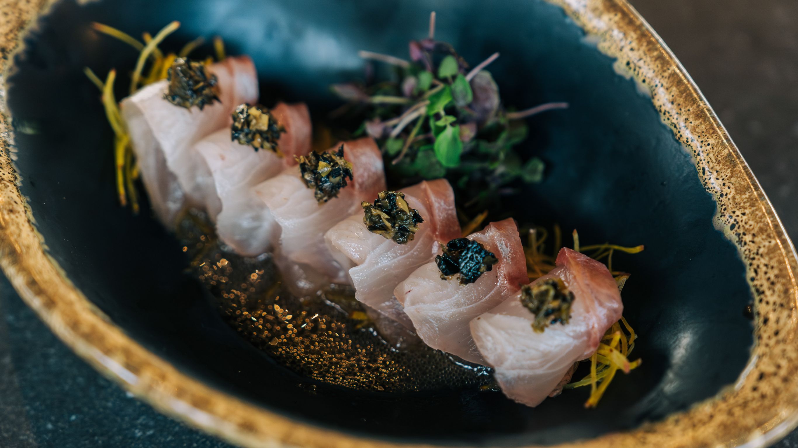 A dish with rolled fish slices topped with caviar served on a dark plate.