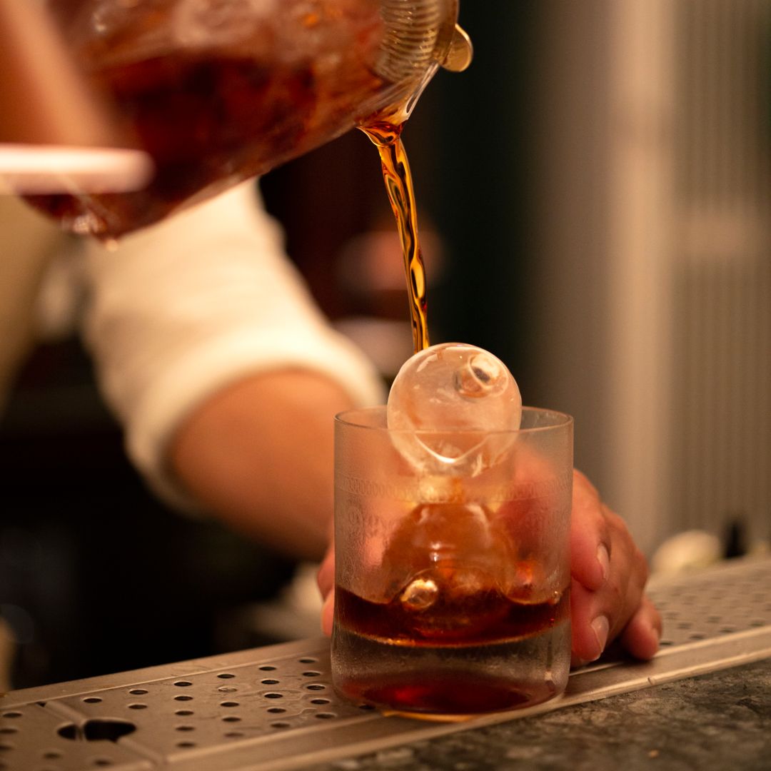 A bartender pouring a drink into a glass with  a large ice cube at a bar.