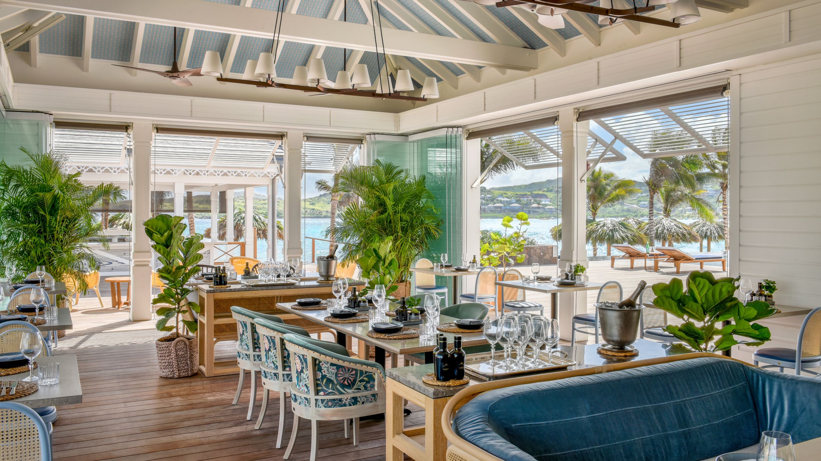 Bright, airy restaurant with large windows, tropical plants, and tables set for dining by the sea.
