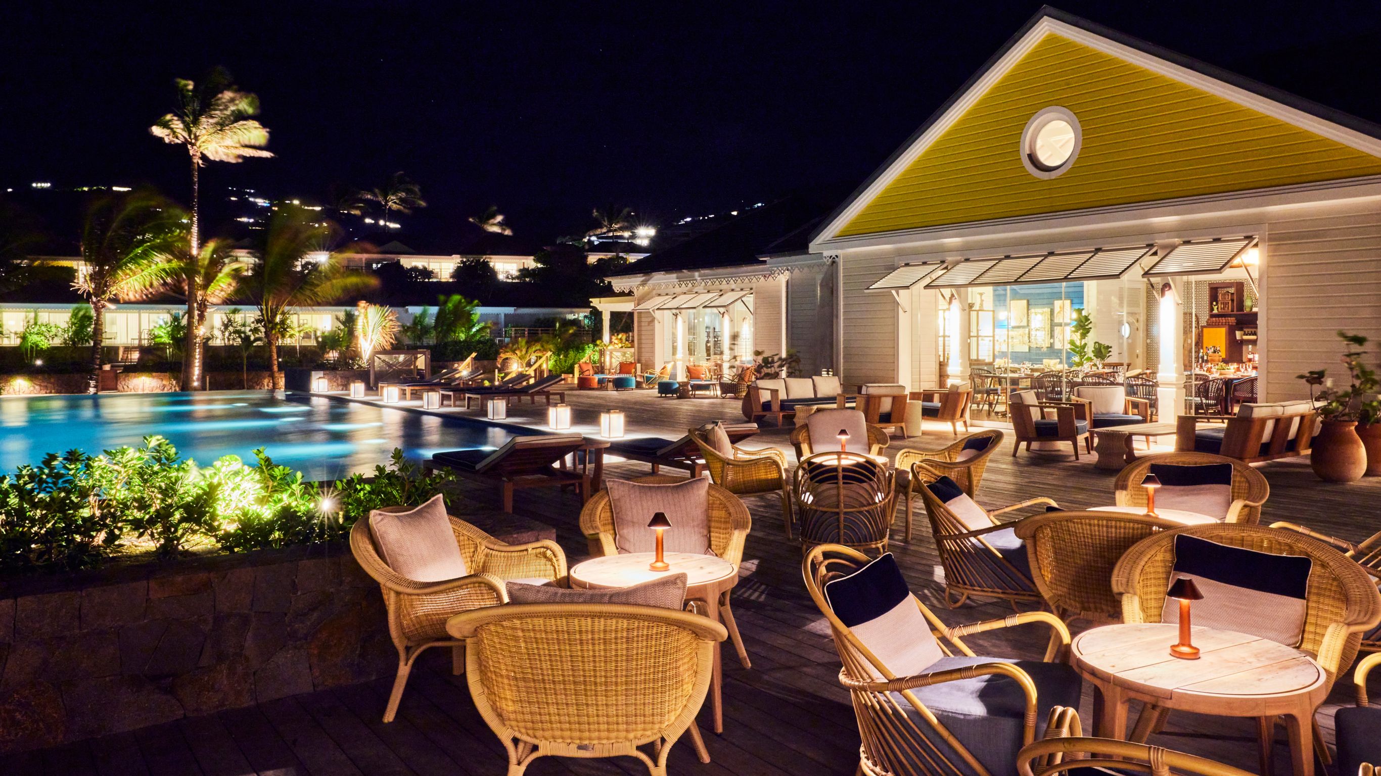 A nighttime view of a poolside setting featuring wicker chairs, tables, and palm trees.