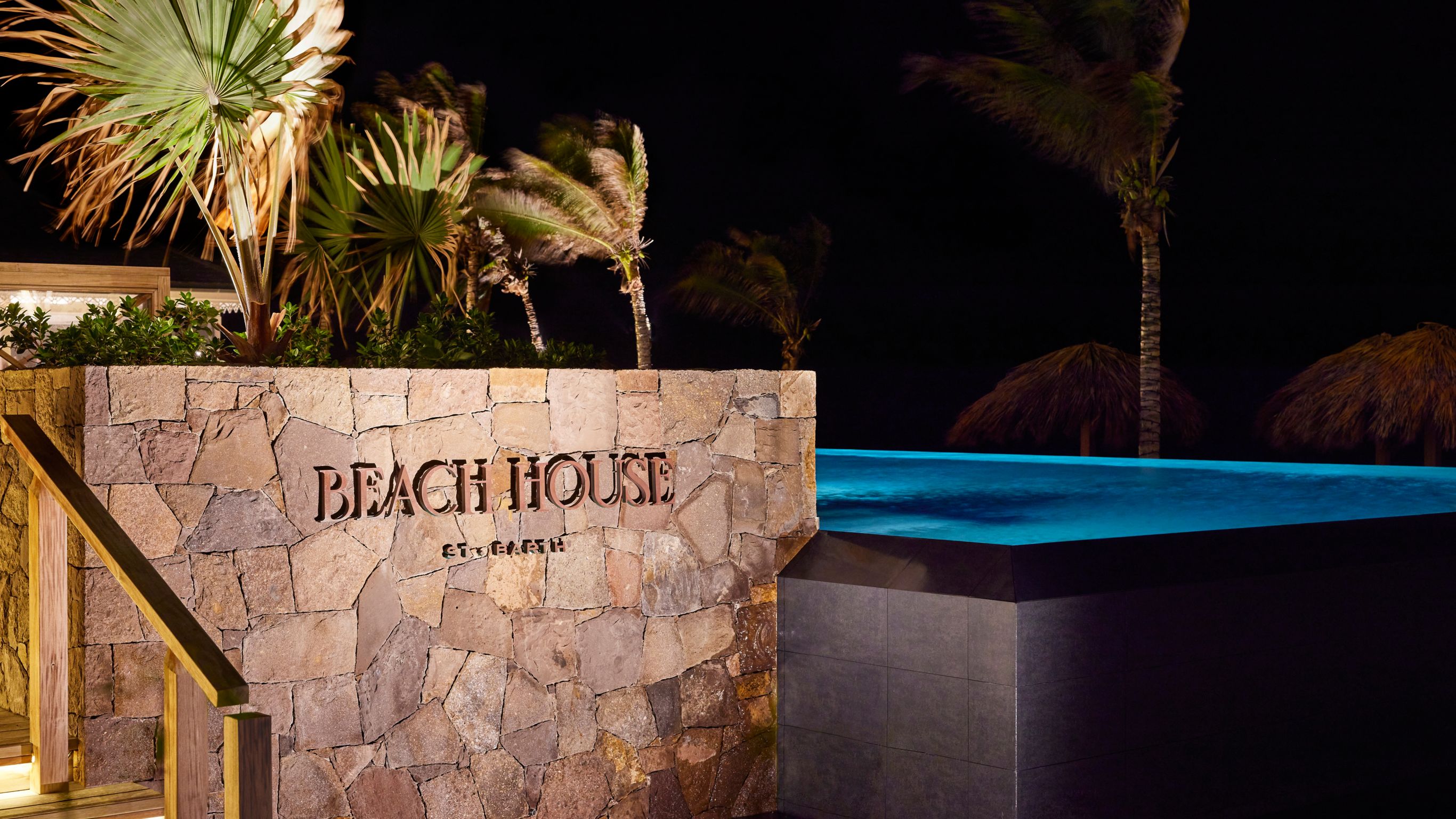 A nighttime view of a pool with a stone wall sign reading "BEACH HOUSE" and palm trees.