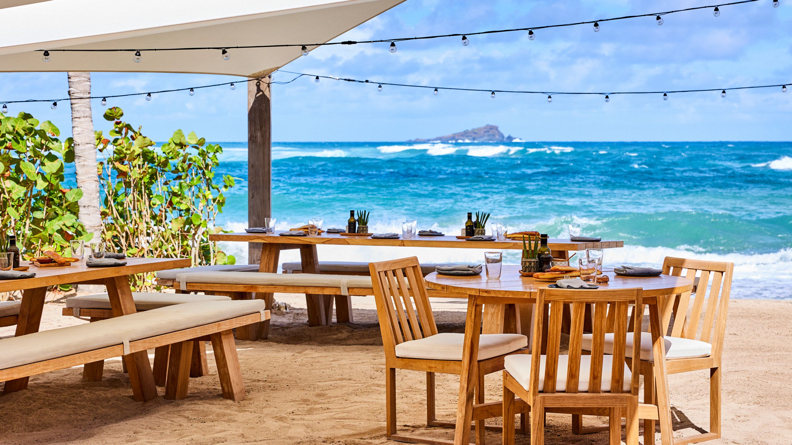 Beachfront dining setup with wooden tables and benches under a canopy, overlooking the ocean.