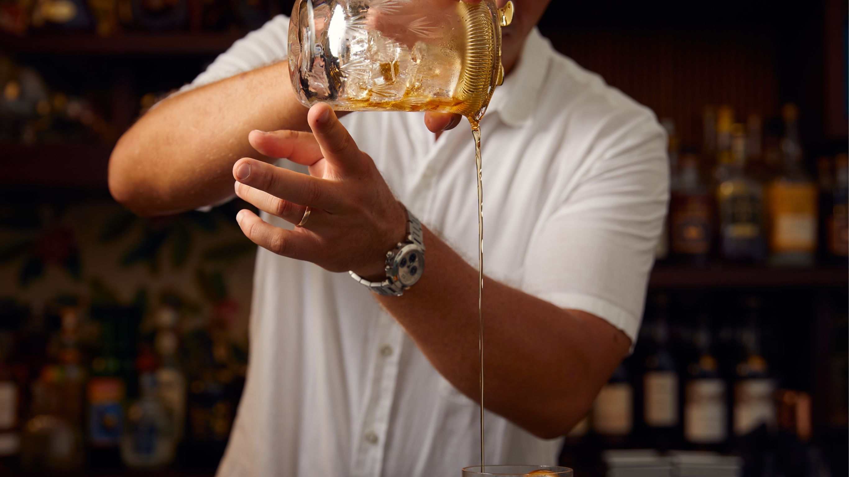 A bartender wearing a white shirt pours a drink from a shaker into a glass.