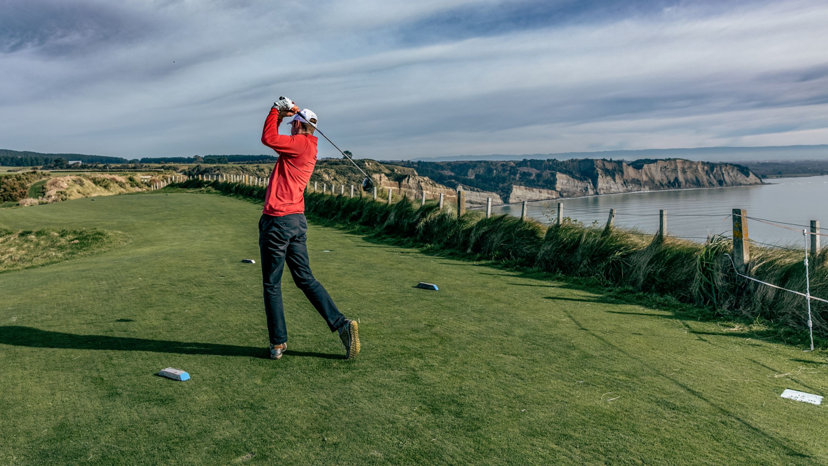 Golfer in a red shirt in mid-swing with a view of cliffs