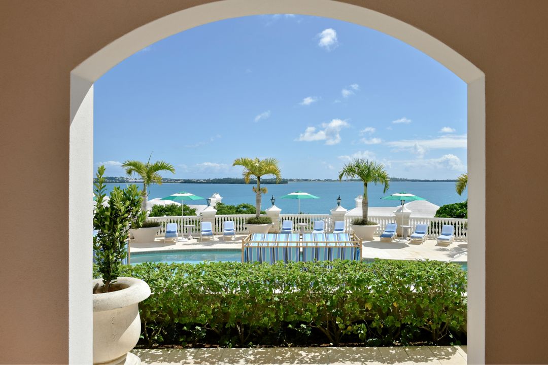 A window overlooking a pool surrounded by greenery and lounge chairs with umbrellas in a Bermuda hotel.