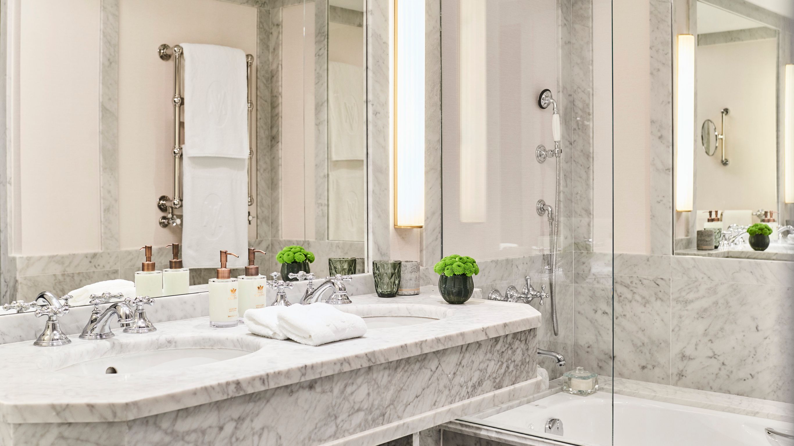 A luxurious marble bathroom with two sinks side by side, multiple hand towels, and a shower with a transparent divider.