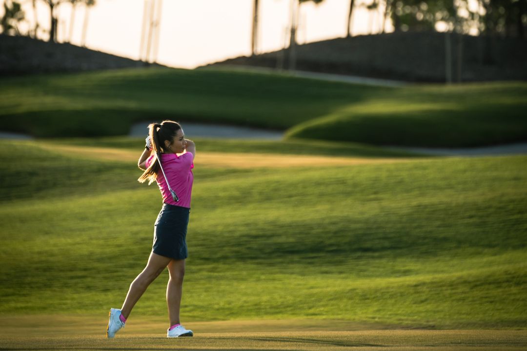 Golfer in mid-swing on a green golf course, with trees and a sunset in the background.
