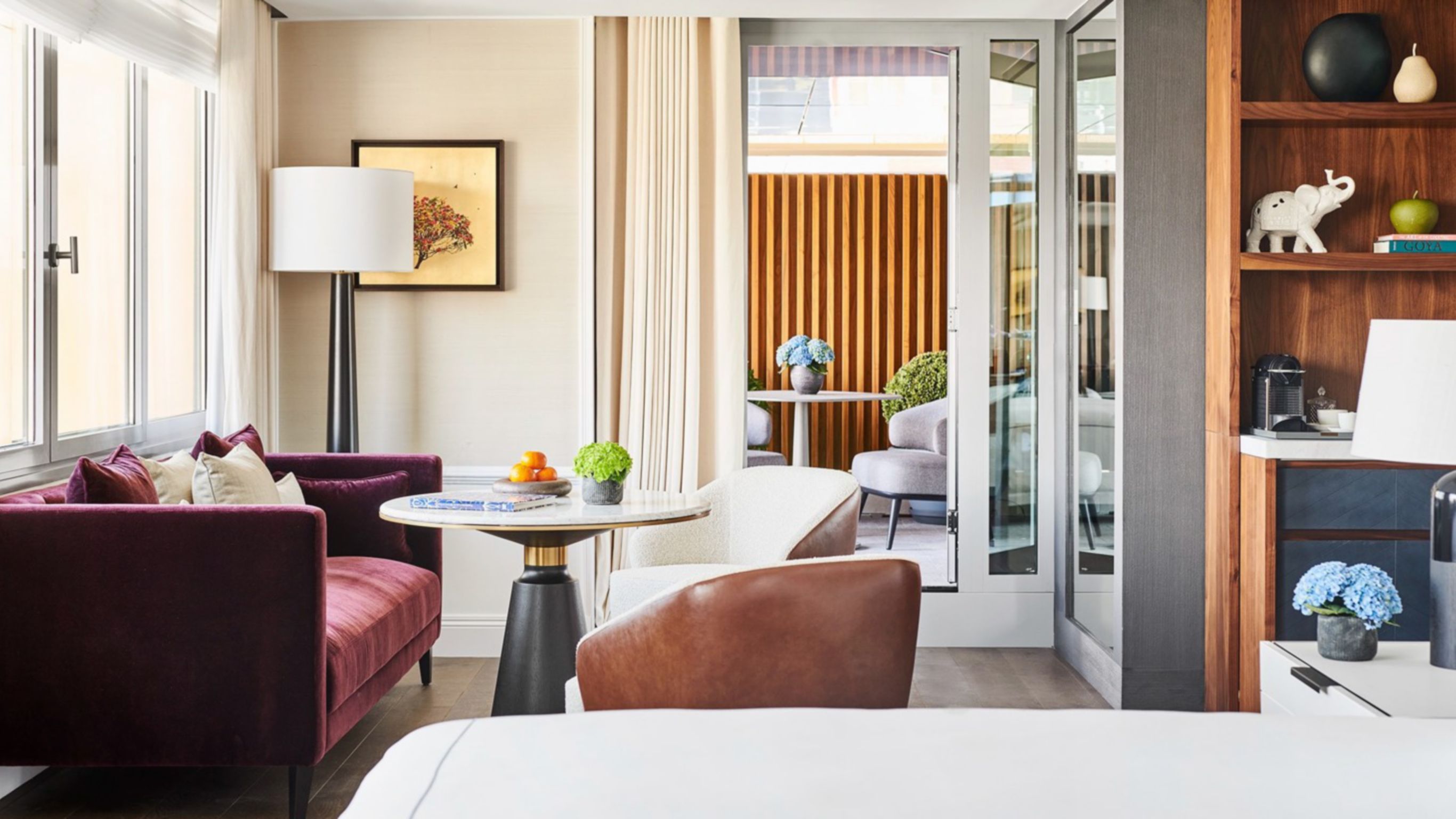 A hotel lounge featuring a burgundy couch, a large lamp, two chairs around a table, and an open sliding door leading to an outdoor table.