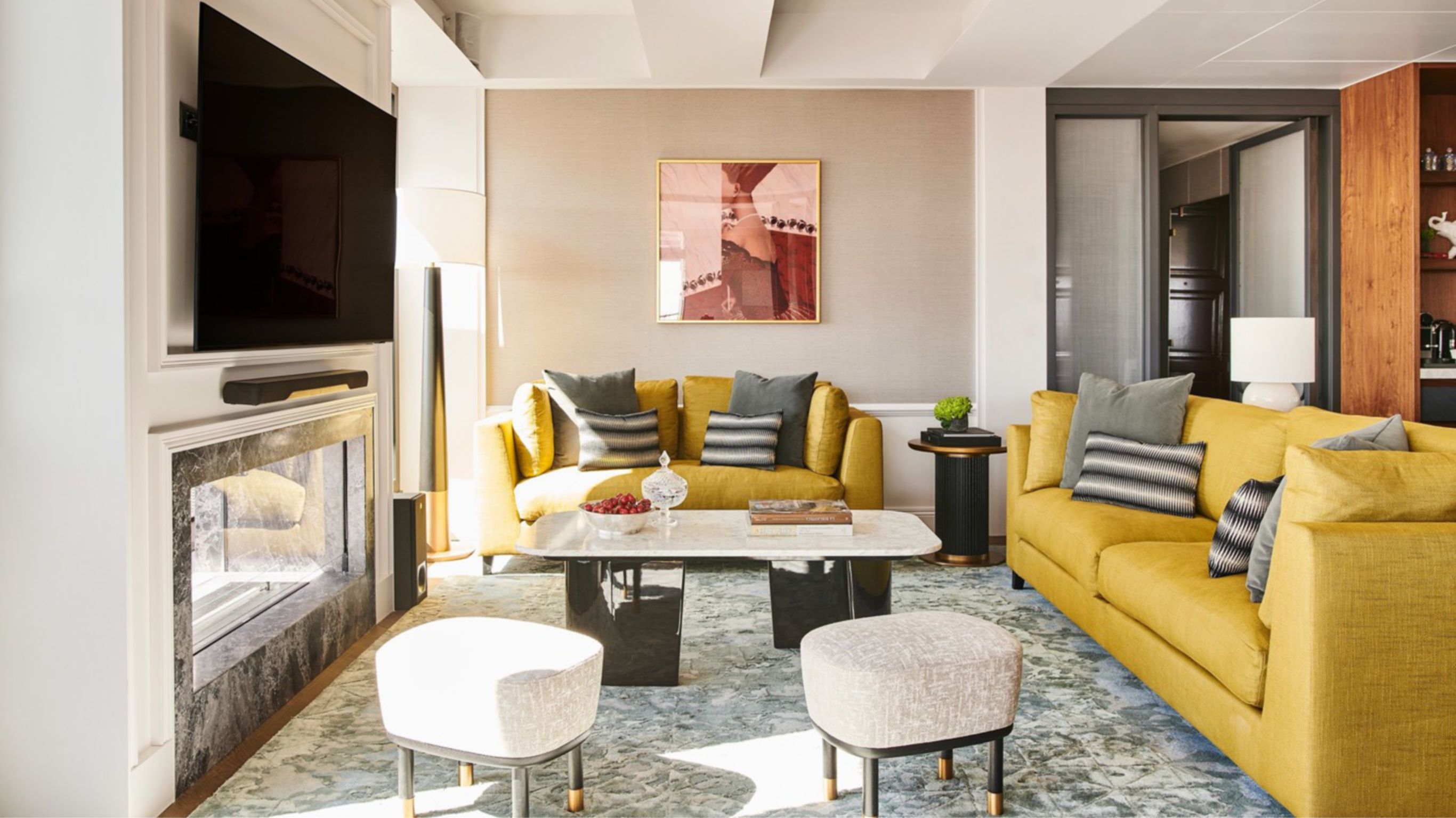 A luxurious sitting area with yellow couches and a coffee table, facing a large flat-screen TV with a fireplace below it.