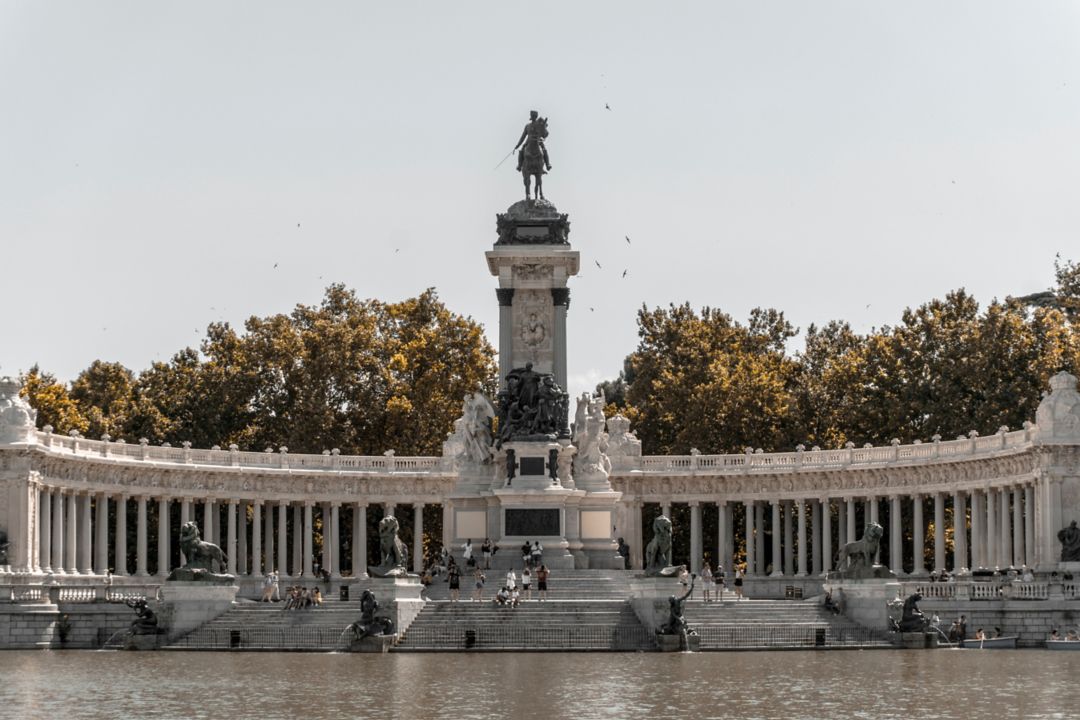 Monument with a statue on top, surrounded by columns and trees with people walking around.
