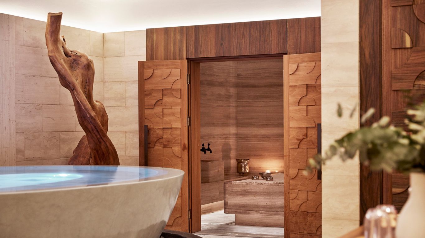 Baño con bañera de piedra, puertas de madera y una escultura de madera, que conduce a una sala de sauna