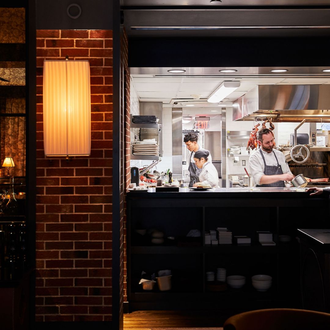 A restaurant kitchen with chefs preparing food, visible through a window from a brick-walled dining area.