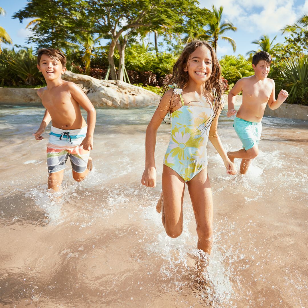 Three children running and playing in shallow water at a beach, with palm trees and greenery.