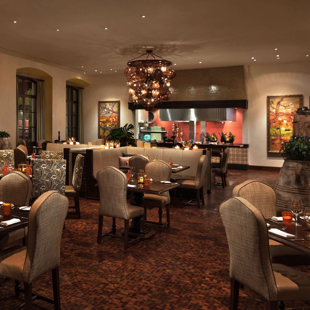 Interior de restaurante con mesas, sillas, un candelabro y una cocina abierta al fondo.