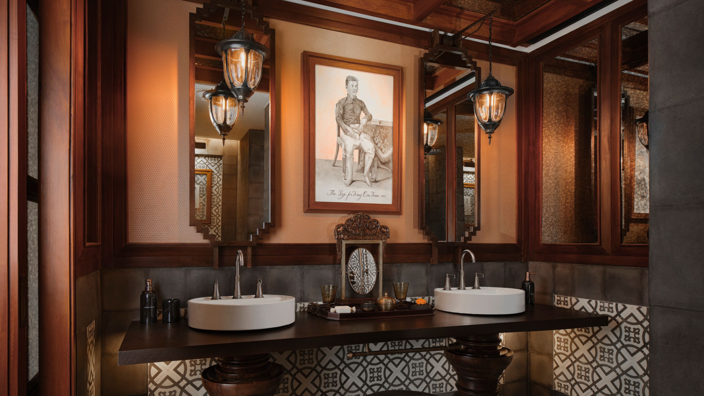 Hotel bathroom designed by Bill Bensley with double sinks, patterned tiles, framed photo, mirrors, and decorative lighting in Luang Prabang