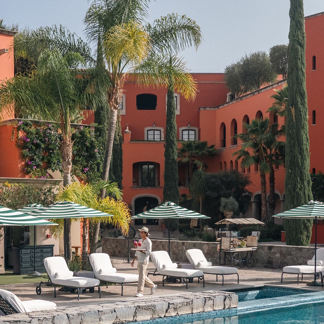 Área de alberca de hotel con camastros bajo toldos, palmeras y toallas enrolladas.