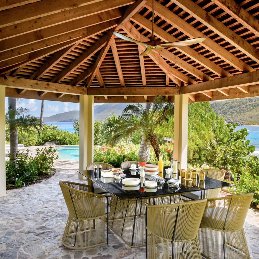 An outdoor dining area with a wooden pergola roof, featuring a table set with dishes and drinks.