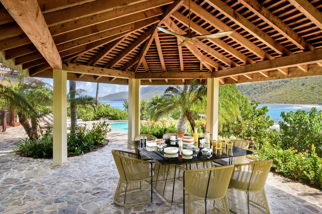 An outdoor dining area with a wooden pergola roof, featuring a table set with dishes and drinks.