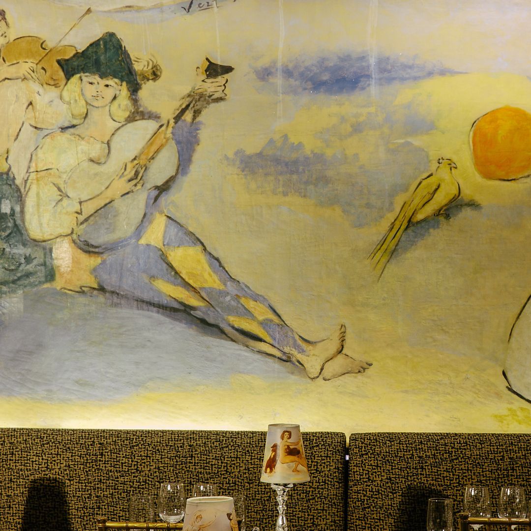 A mural with figures and a bird, above a table with glassware and a decorative lamp.