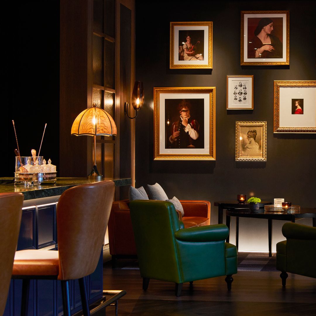 Dimly lit bar with framed artwork on the walls, leather seating, a bar counter, and a lamp.