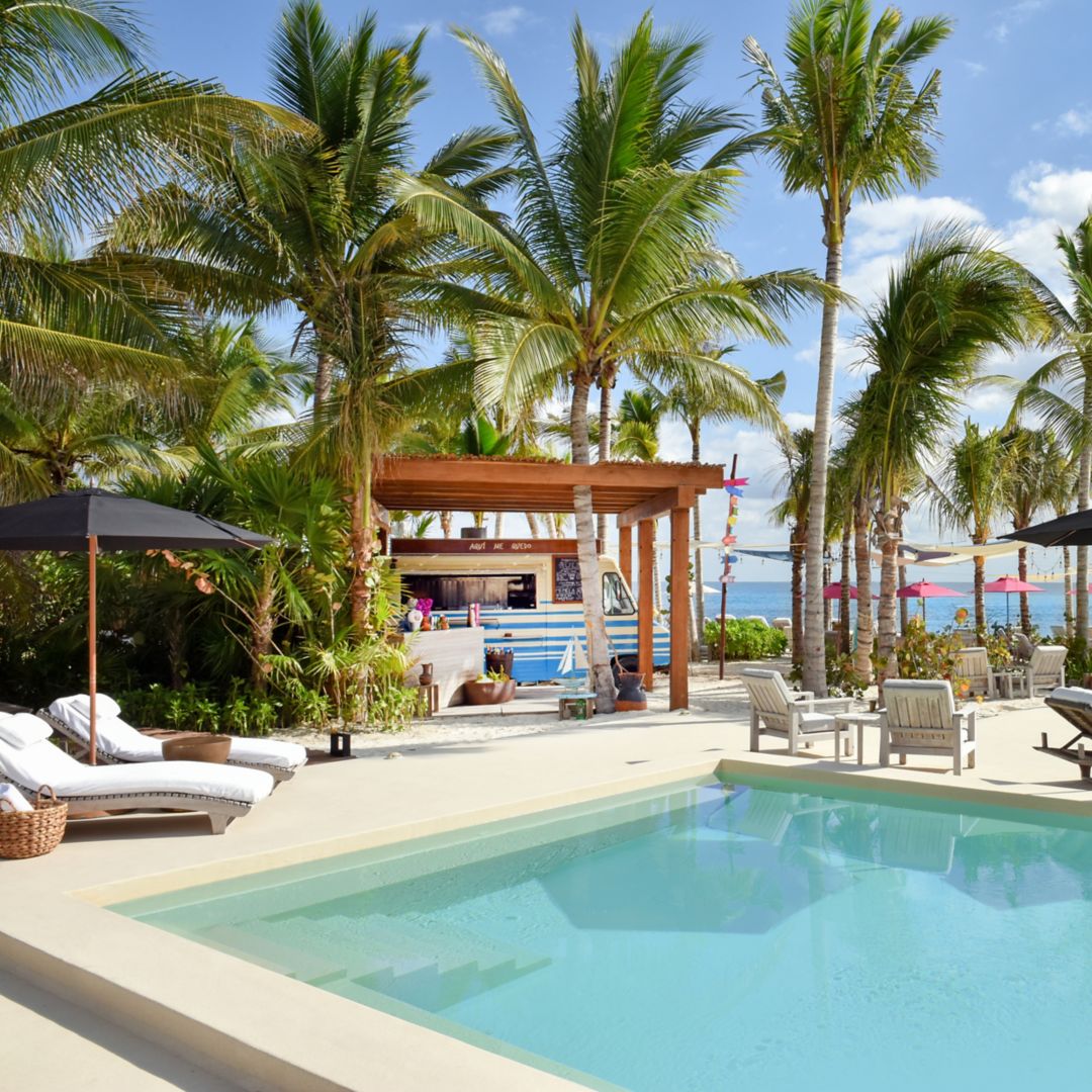 Image Rosewood Mayakoba's Aquí Me Quedo Pools: pictured two plunge pools sunken into the ssan facing the ocean. Surrounded by palm trees, white lounge chairs, blue umbrellas, and food truck.