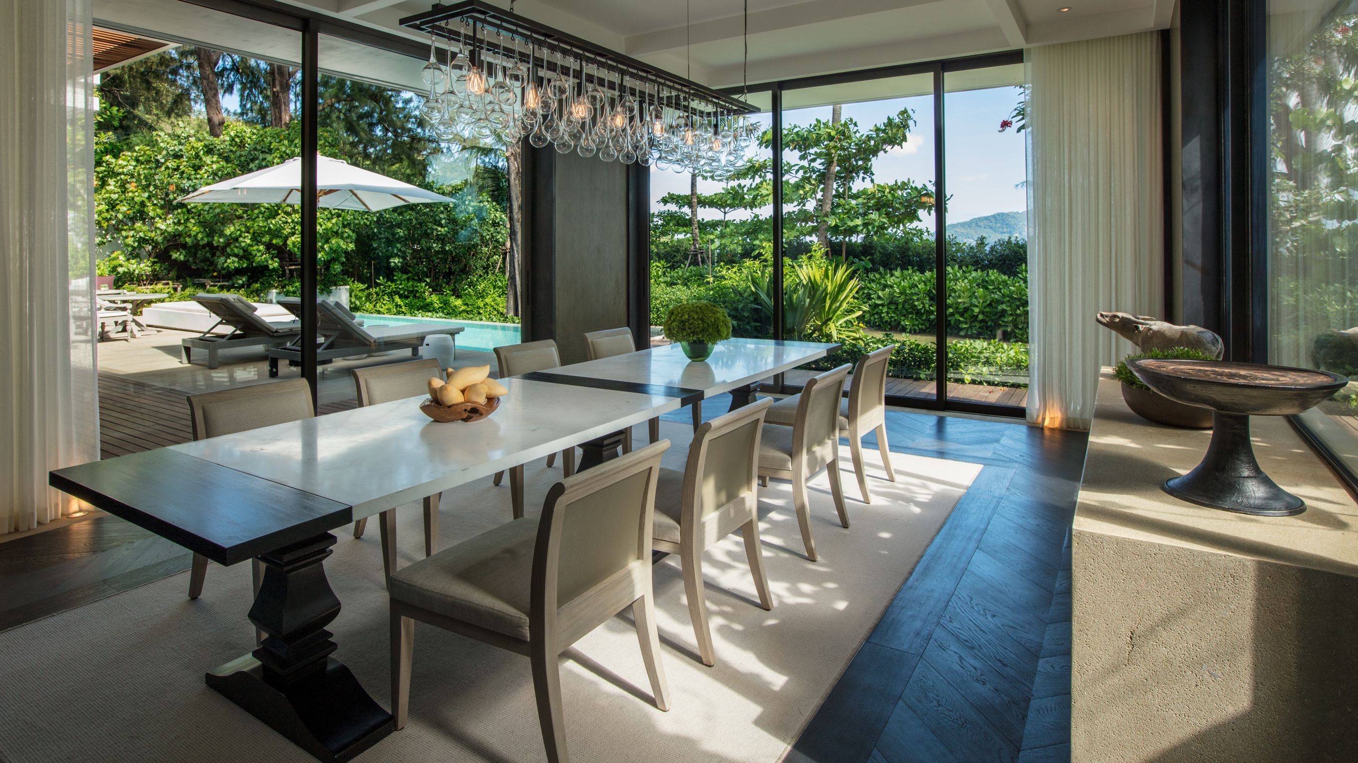 A private villa dining room with a large table, gray chairs, a table on the right side and a pool outside.