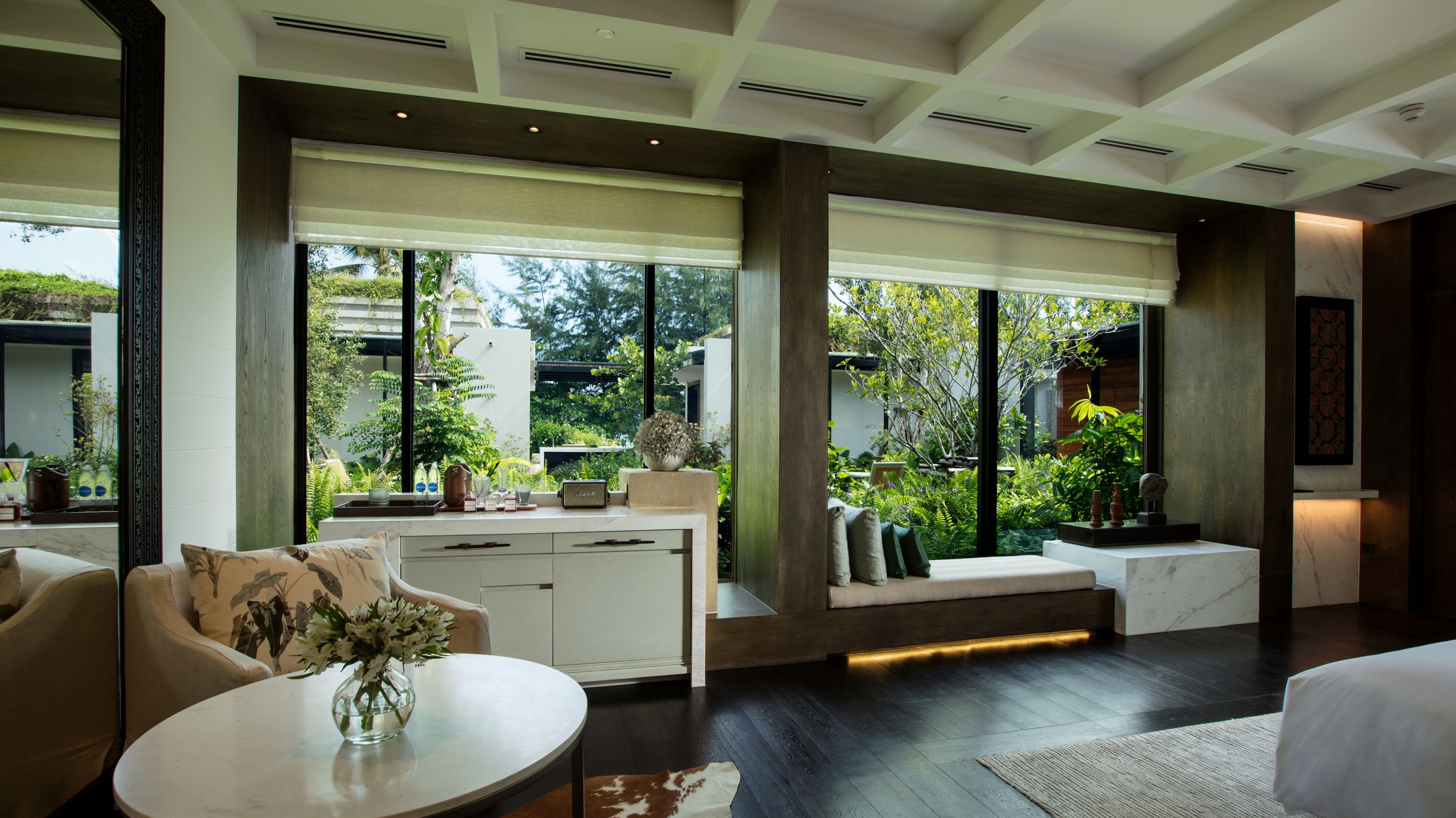 A partial view of a hotel suite, with a coffee table next to a beige chair, drawers, and large windows with trees outside.