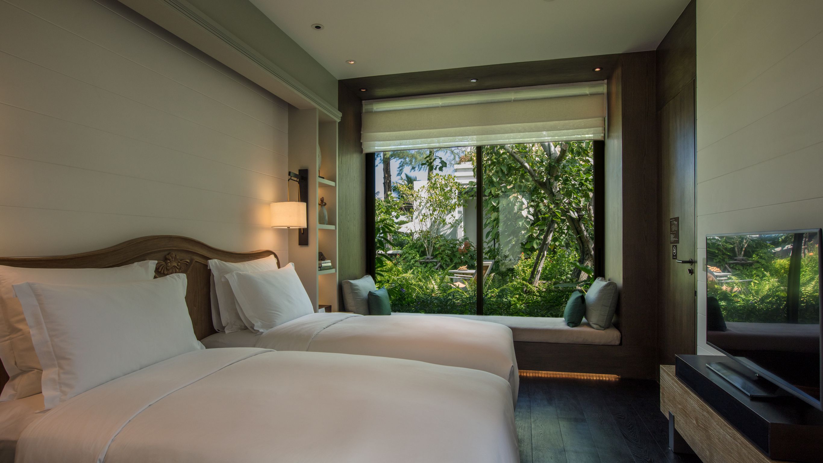 A hotel room with two single beds, shelves with decorations, dim lights, and windows showing trees outside.